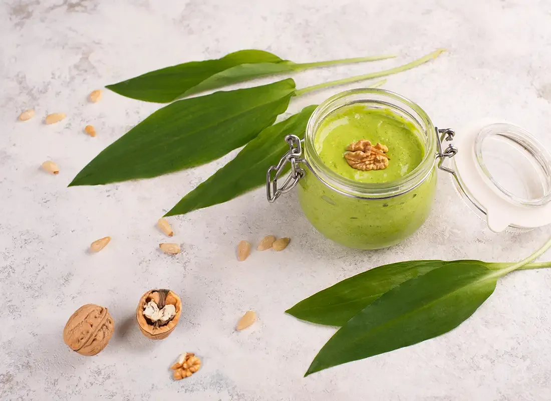 Bärlauchpesto im Glas mit Walnuss, frischen Blättern und Pinienkernen auf hellem Untergrund