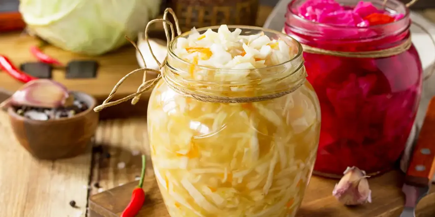 Zwei Einmachgläser mit fermentiertem Weiß- und Rotkraut, dekorativ auf einem Holzbrett mit Gewürzen und Chili angerichtet.