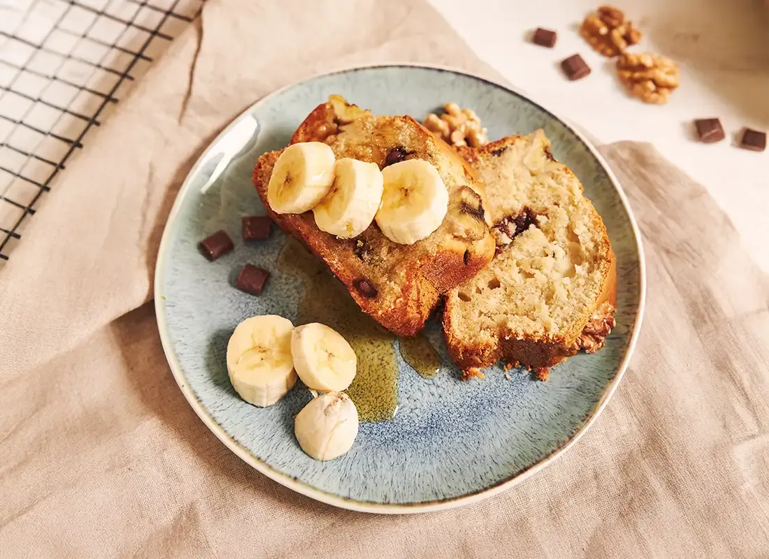 Frisches Bananenbrot mit Schokoladenstückchen, garniert mit Bananenscheiben auf einem blauen Teller, daneben Walnüsse und Schokodrops.