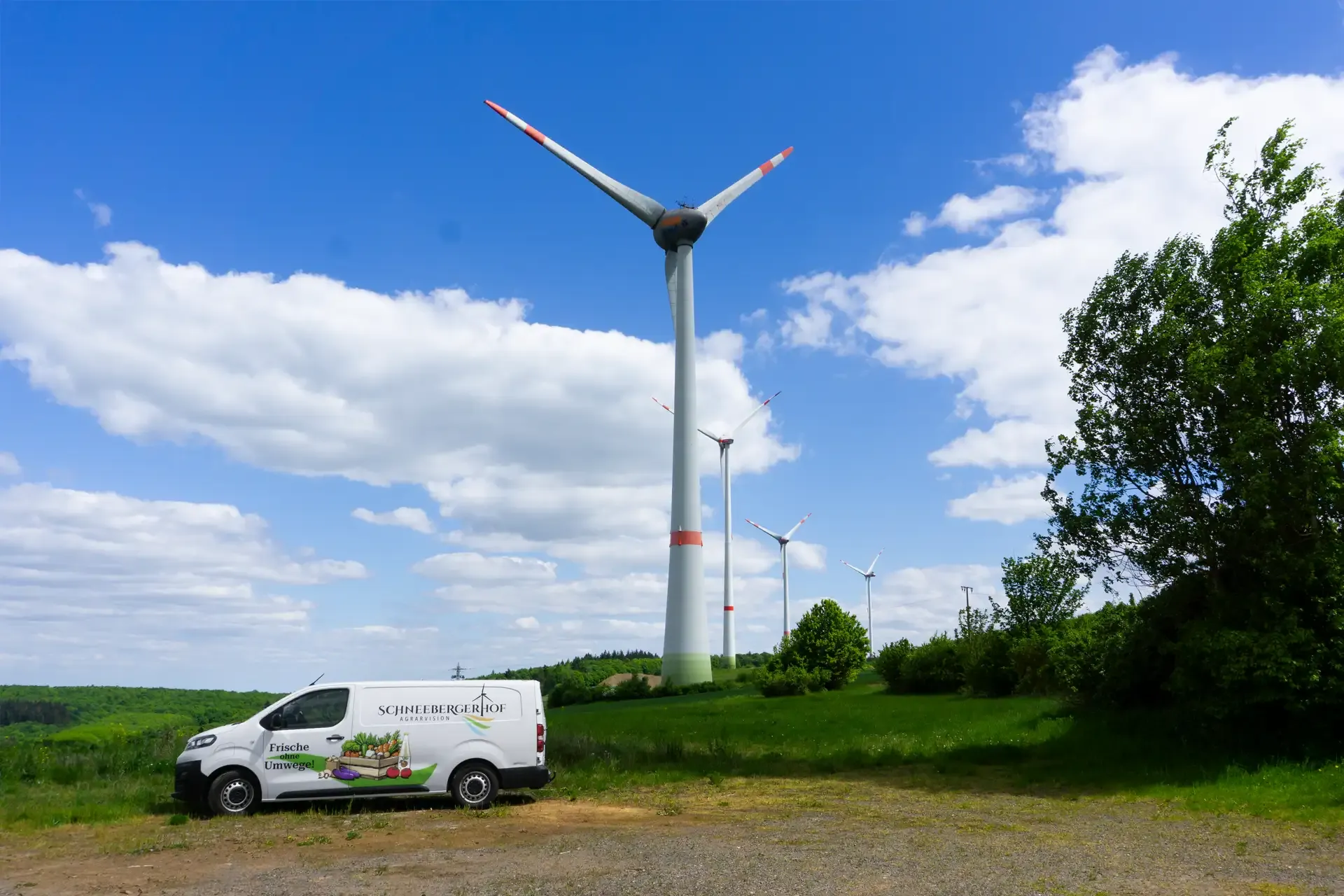 E-Transporter vom Schneebergerhof vor hofeigenen Windrädern auf weiter Wiese unter blauem Himmel