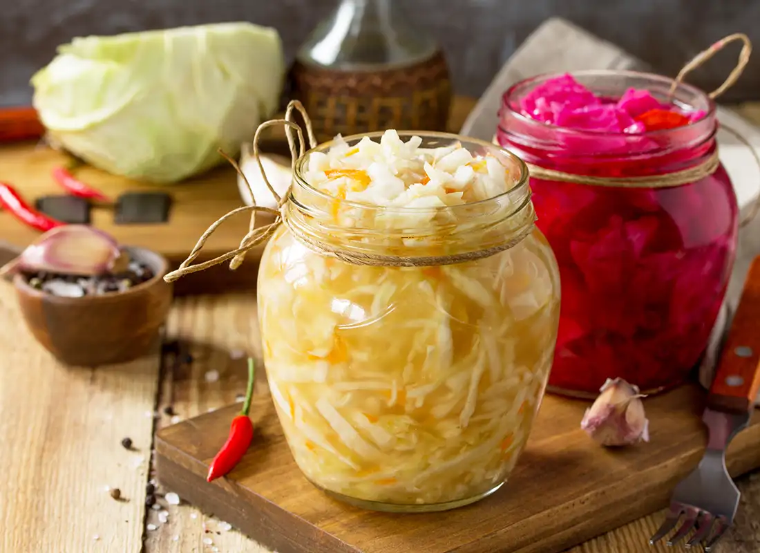 Zwei Einmachgläser mit fermentiertem Weiß- und Rotkraut, dekorativ auf einem Holzbrett mit Gewürzen und Chili angerichtet.